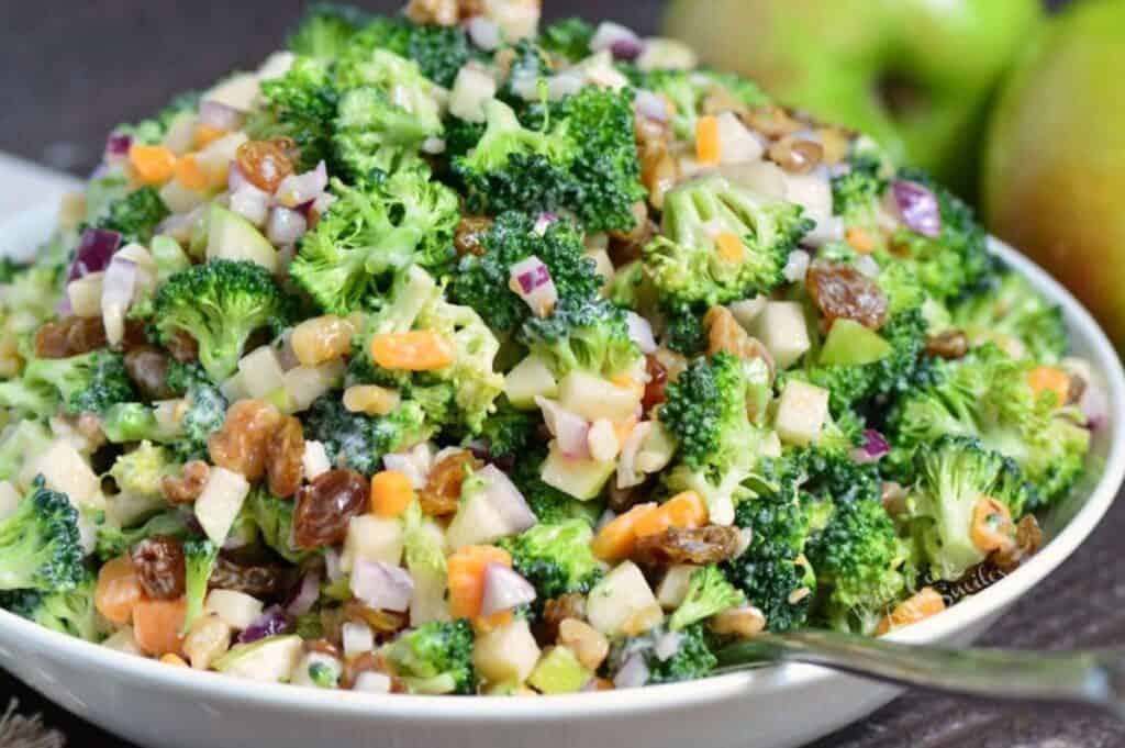 A colorful salad consisting of fresh broccoli florets, chopped red onions, diced apples, raisins, and nuts, all mixed with a creamy dressing, served in a white bowl with a spoon. In the background, there are green apples.
