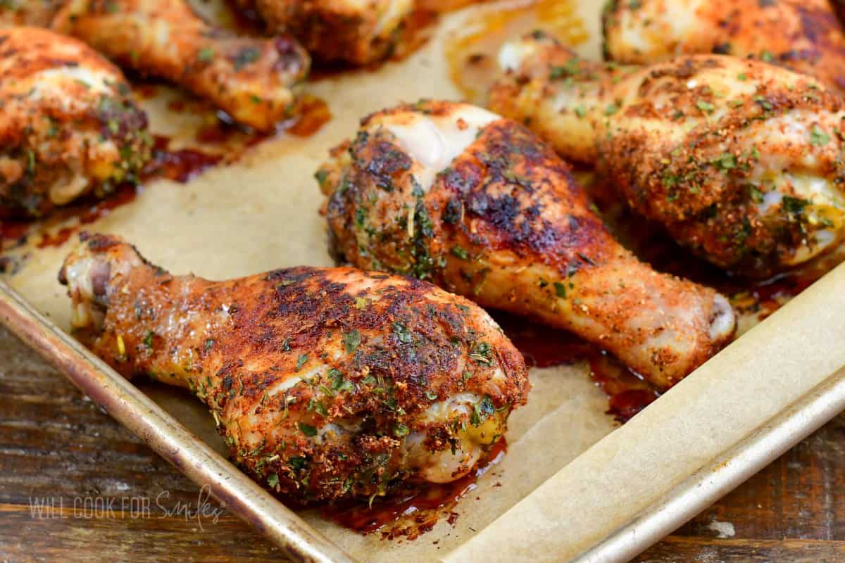 Baked chicken legs are spread across a parchment lined baking sheet.