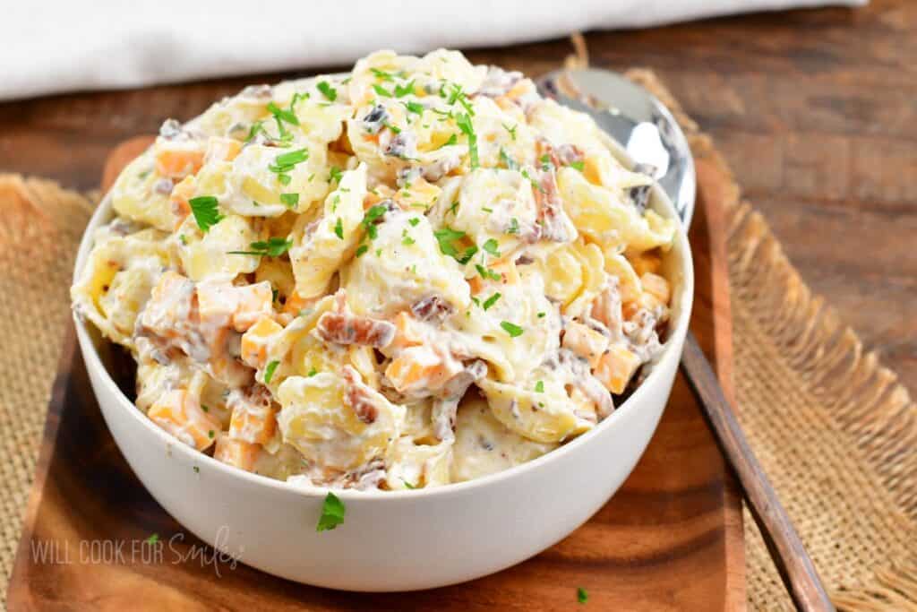 A white bowl filled with a creamy pasta salad, featuring a mix of shell pasta, bacon pieces, and diced vegetables, garnished with fresh parsley. A spoon rests inside the bowl, which sits on a wooden cutting board. A burlap cloth is partially visible underneath.