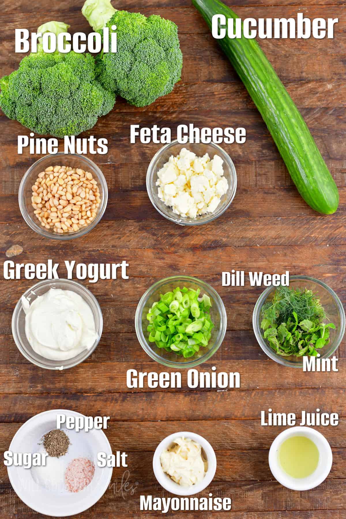 Labeled ingredients for broccoli cucumber salad are placed on a wooden surface. 