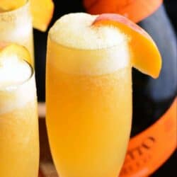a champagne flute filled with orange peach Bellini cocktail with more on background.