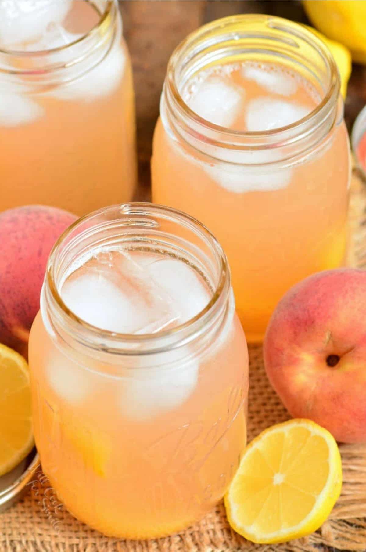 three mason jars filled with peach lemonade and ice with peaches around.