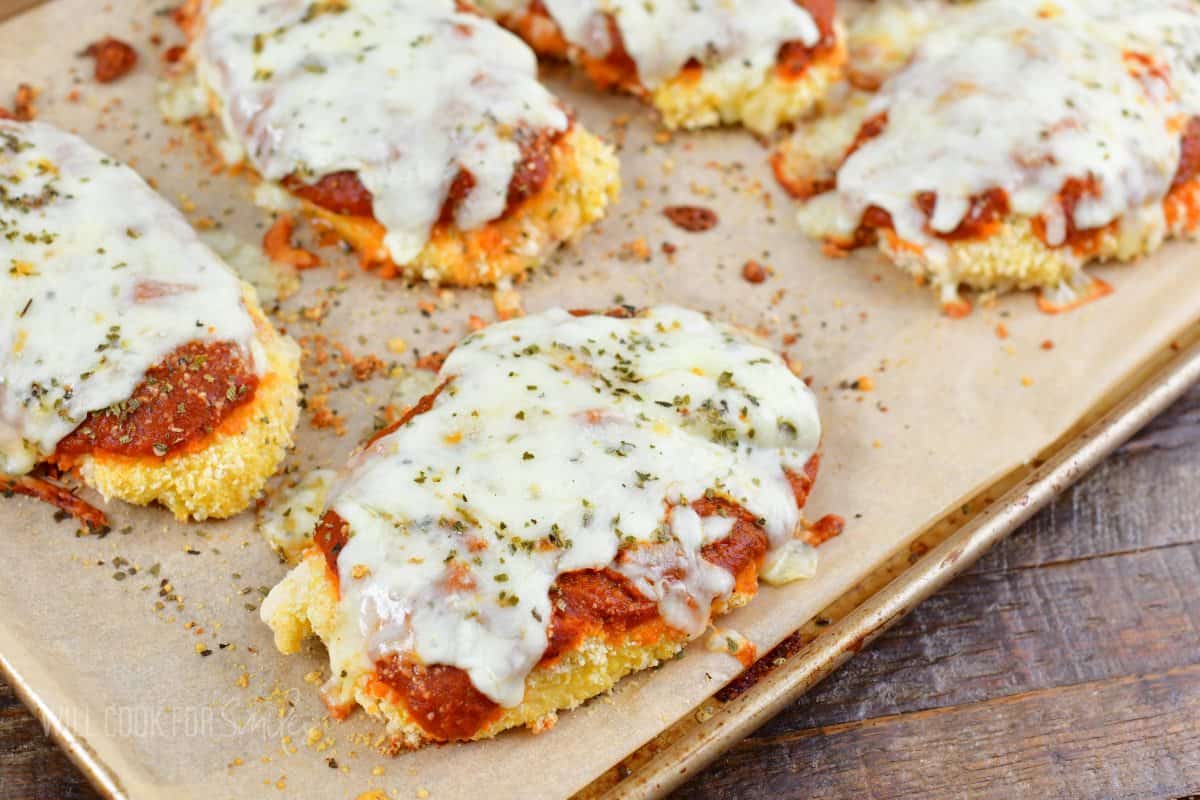 several pieces of baked chicken parmesan on baking sheet with sauce and melted cheese.