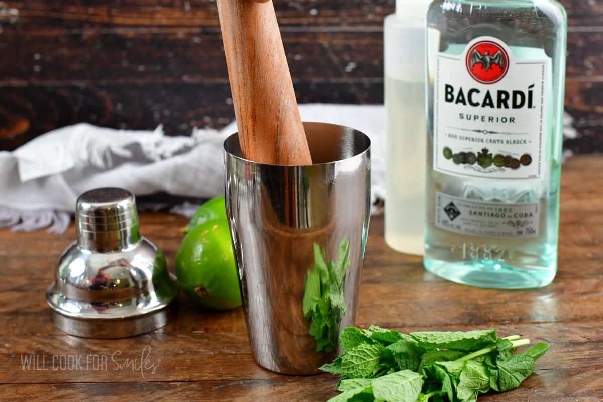 muddling mint in the silver cocktail shaker next to rum and mint on the table.