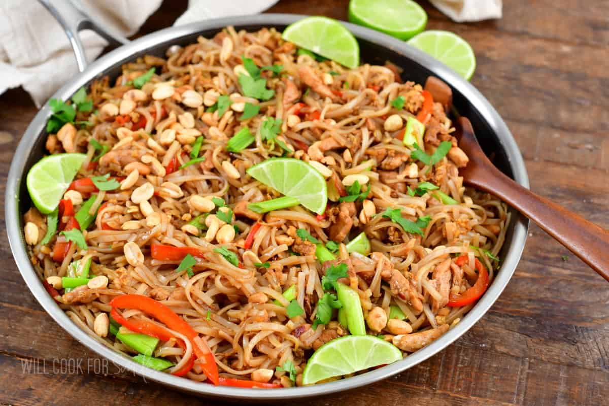 A wooden spoon is placed in a skillet filled with garnished pad Thai. 