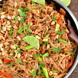 pad thai in a pan with lime wedges on a wood surface.