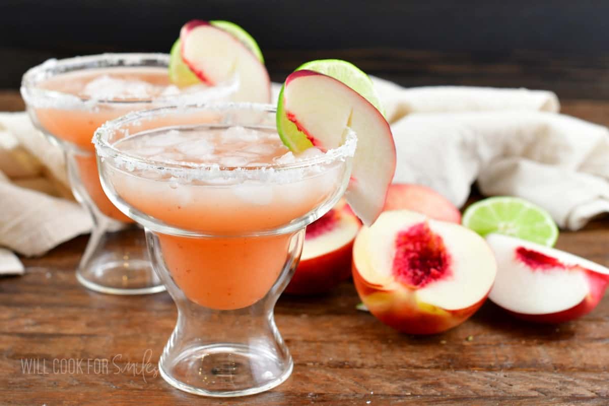 Two garnished margaritas are placed next to sliced peaches.