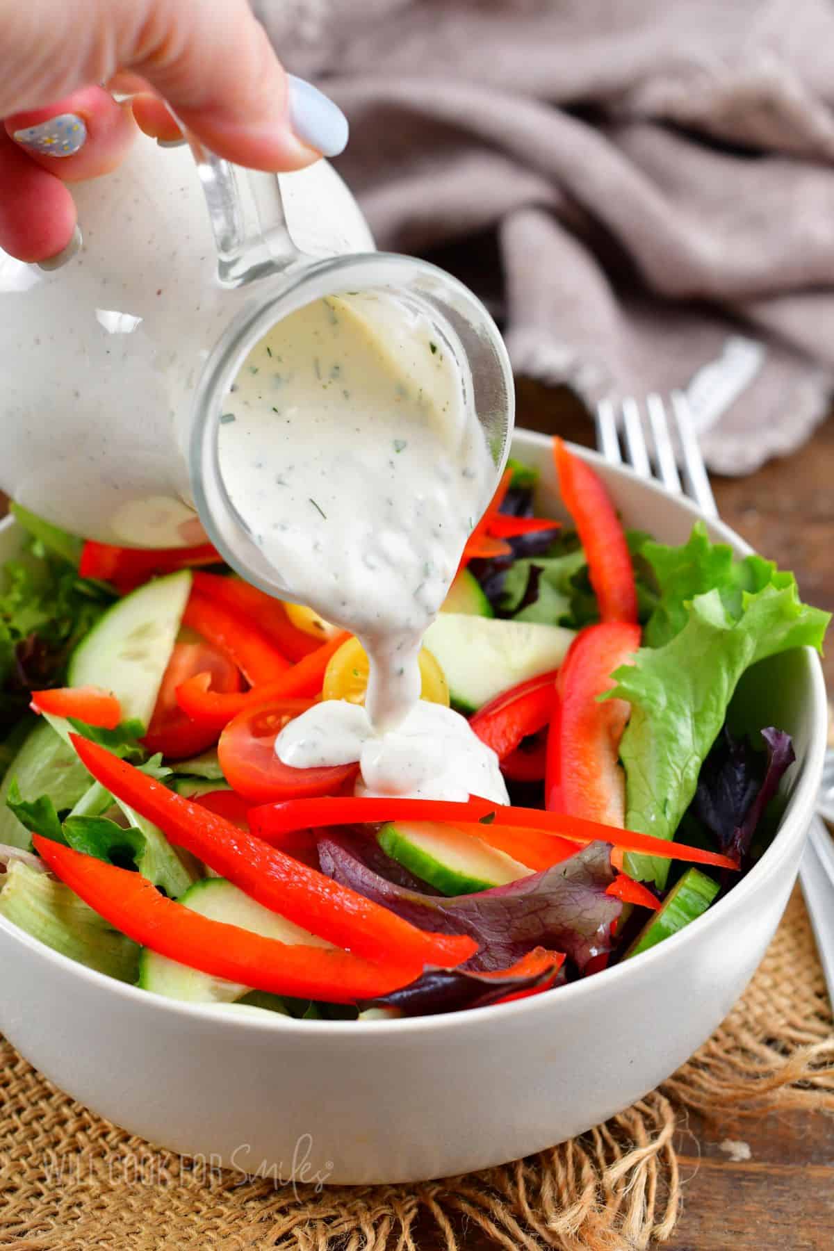 pouring ranch dressings over a green salad with peppers and cucumber.