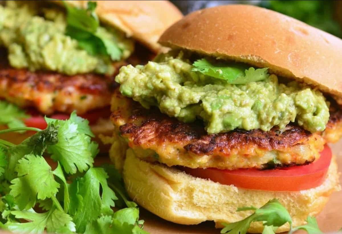 Two fully assembled shrimp burgers are placed next to a bunch of cilantro. 