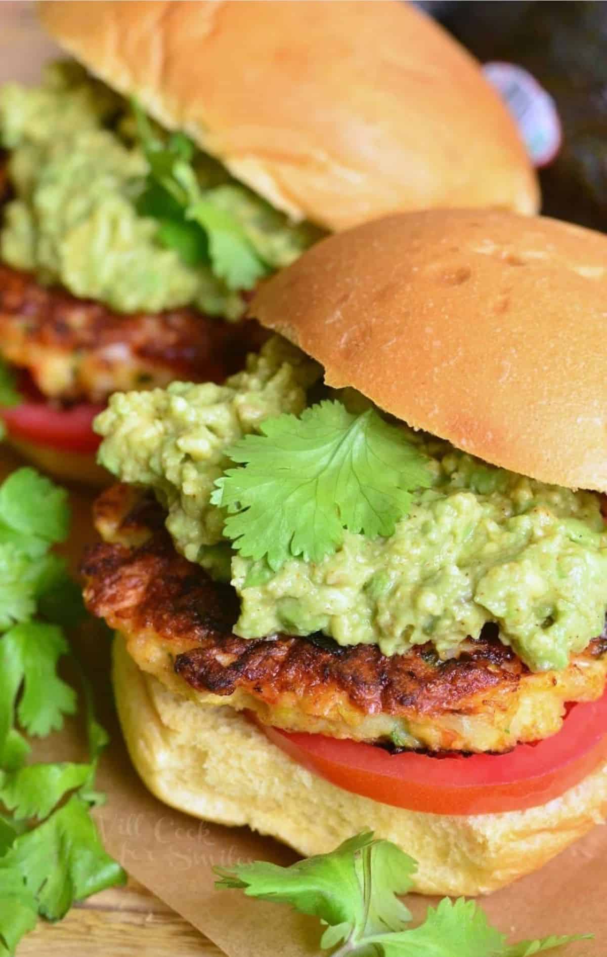 two assembled shrimp burgers with guacamole and tomato on a bun. 
