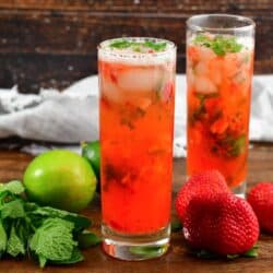 two tall cocktails with bright strawberry mojitos, and berries, lime and mint around.