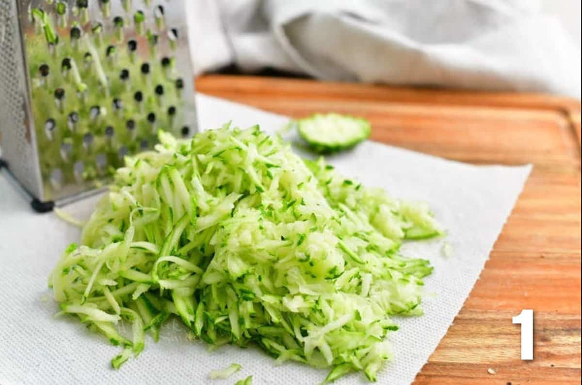 Shredded zucchini is placed on a paper towel next to a cheese grater. 