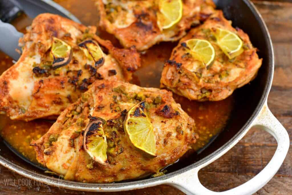A pan filled with four pieces of seasoned and roasted chicken topped with sliced limes. The chicken appears to be well-cooked and browned, with visible herbs and spices. The dish is set on a wooden surface.
