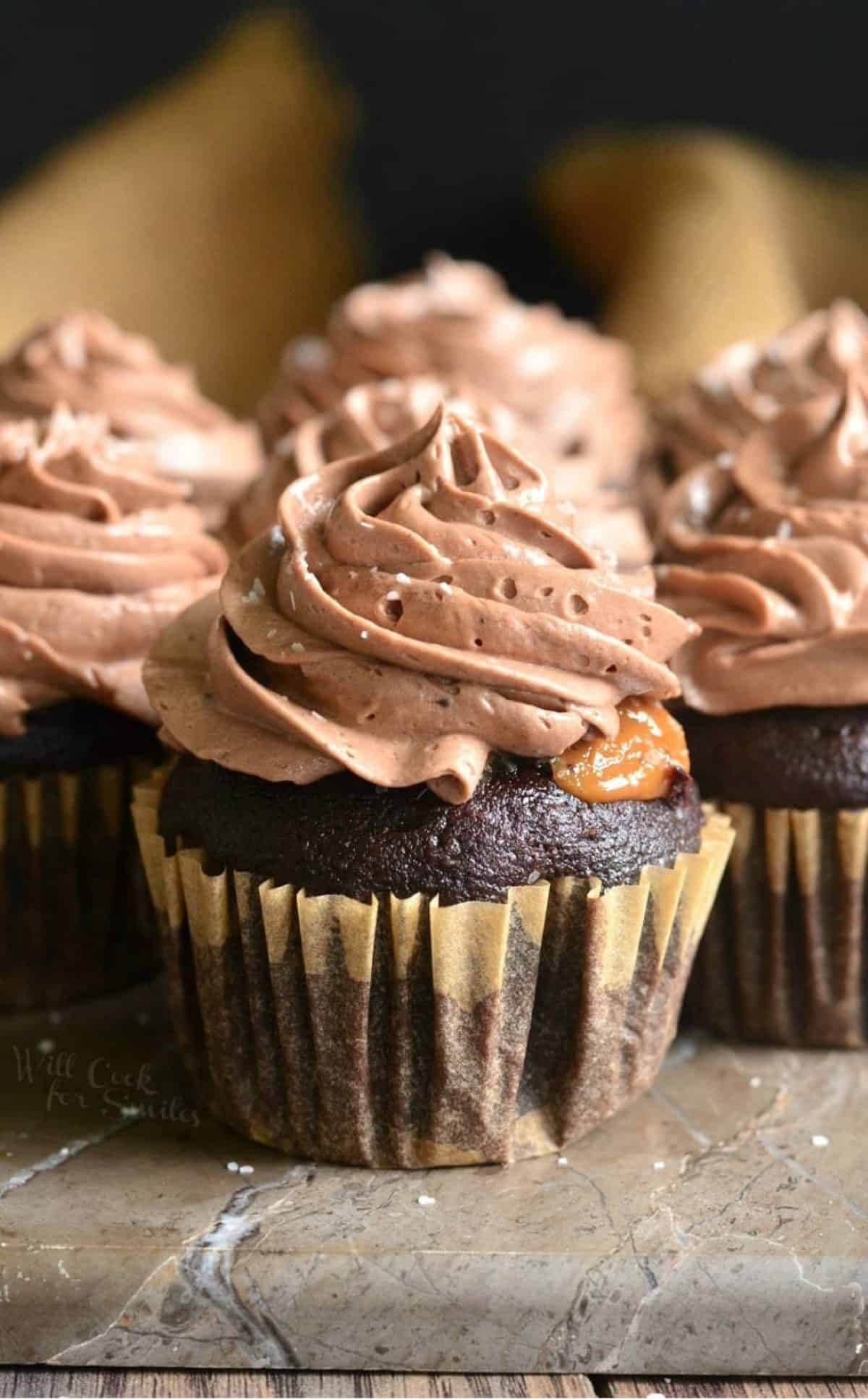 A Chocolate cupcake with chocolate frosting and salted dulce de leche filling sitting on the foreground and more behind.