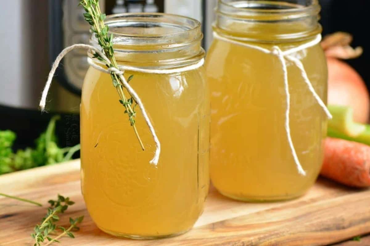 two mason jars of stock with thyme tied to one and twine tied around the other on a wood surface.