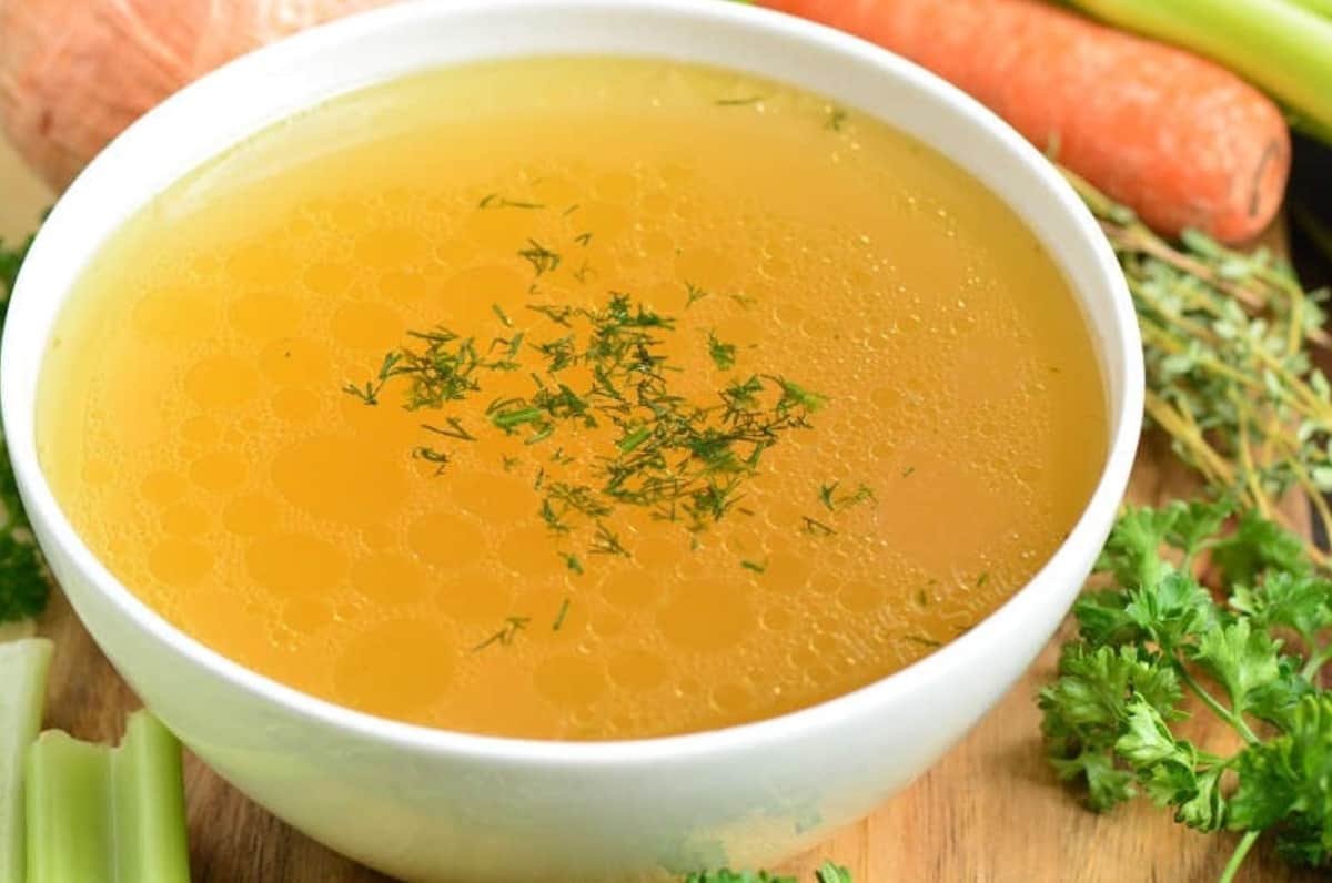 bowl of chicken stock with herbs on top as garnish and carrot, celery, and herbs on a wood surface.