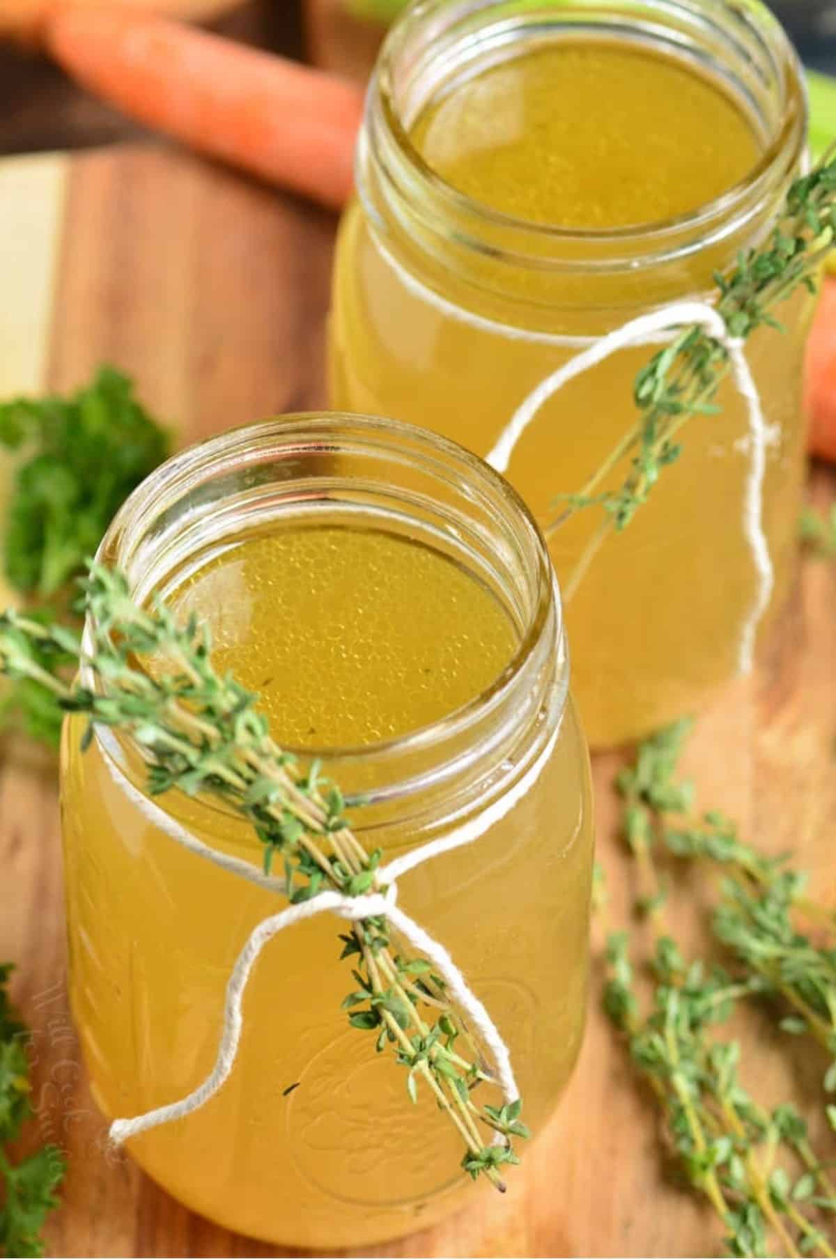 two mason jars of chicken stock with thyme tied to around them on a wood surface.