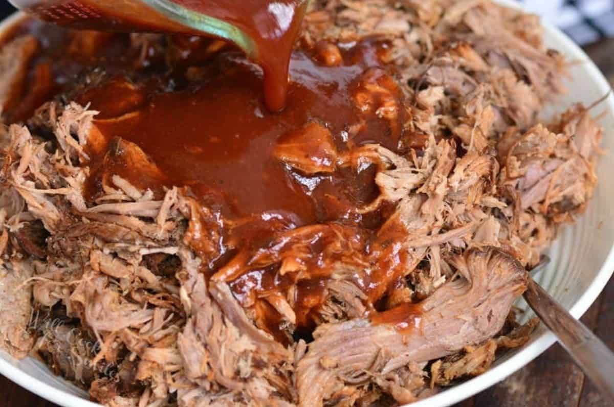 pouring bbq sauce over pulled pork that is in a white bowl.
