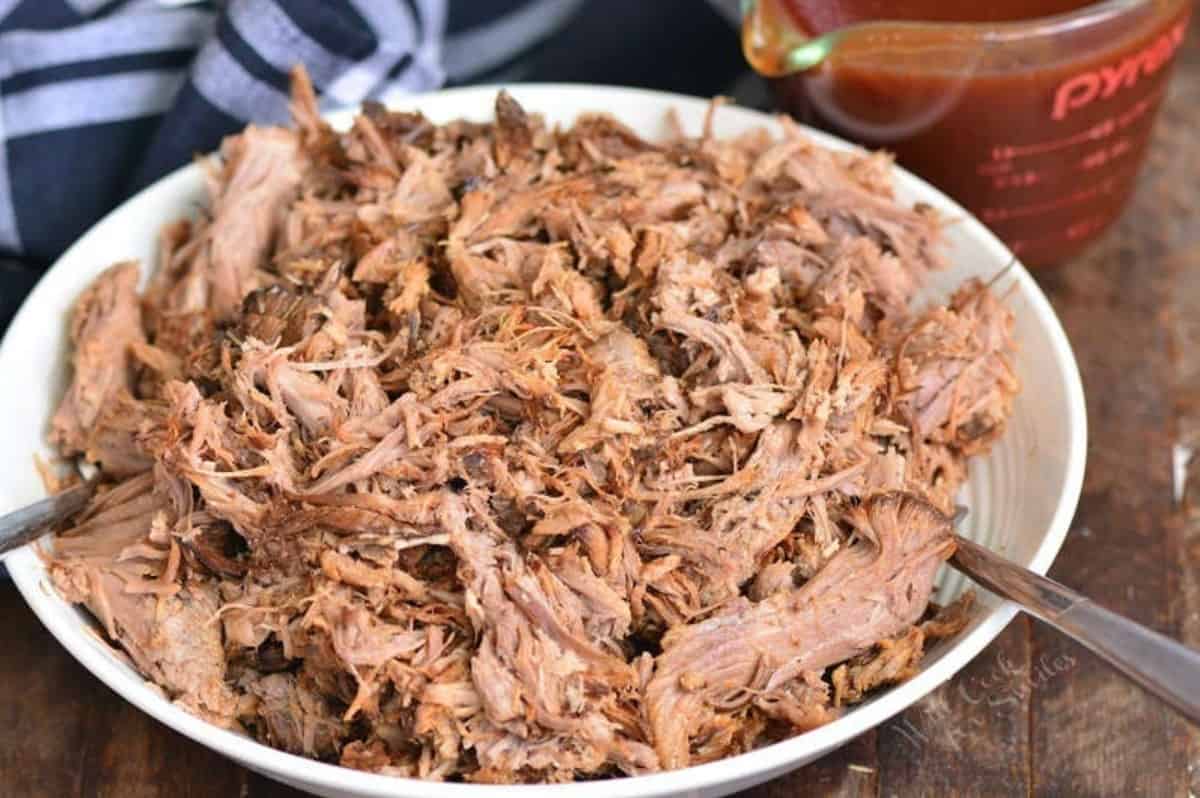 Pulled pork in a white bowl with forks on each side and bbq sauce in a glass measuring cup.