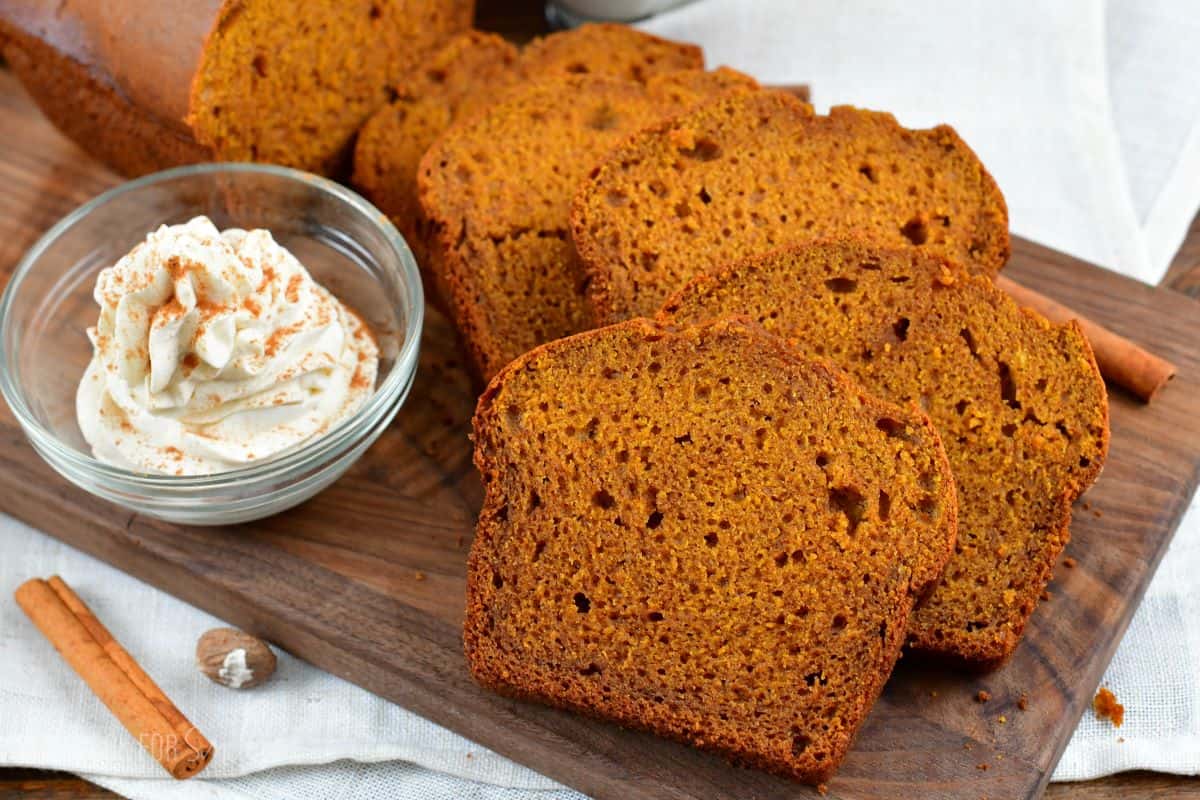slices of pumpkin bread on a wood surface with a bowl of whipped cream with pumpkin spice sprinkled on top.