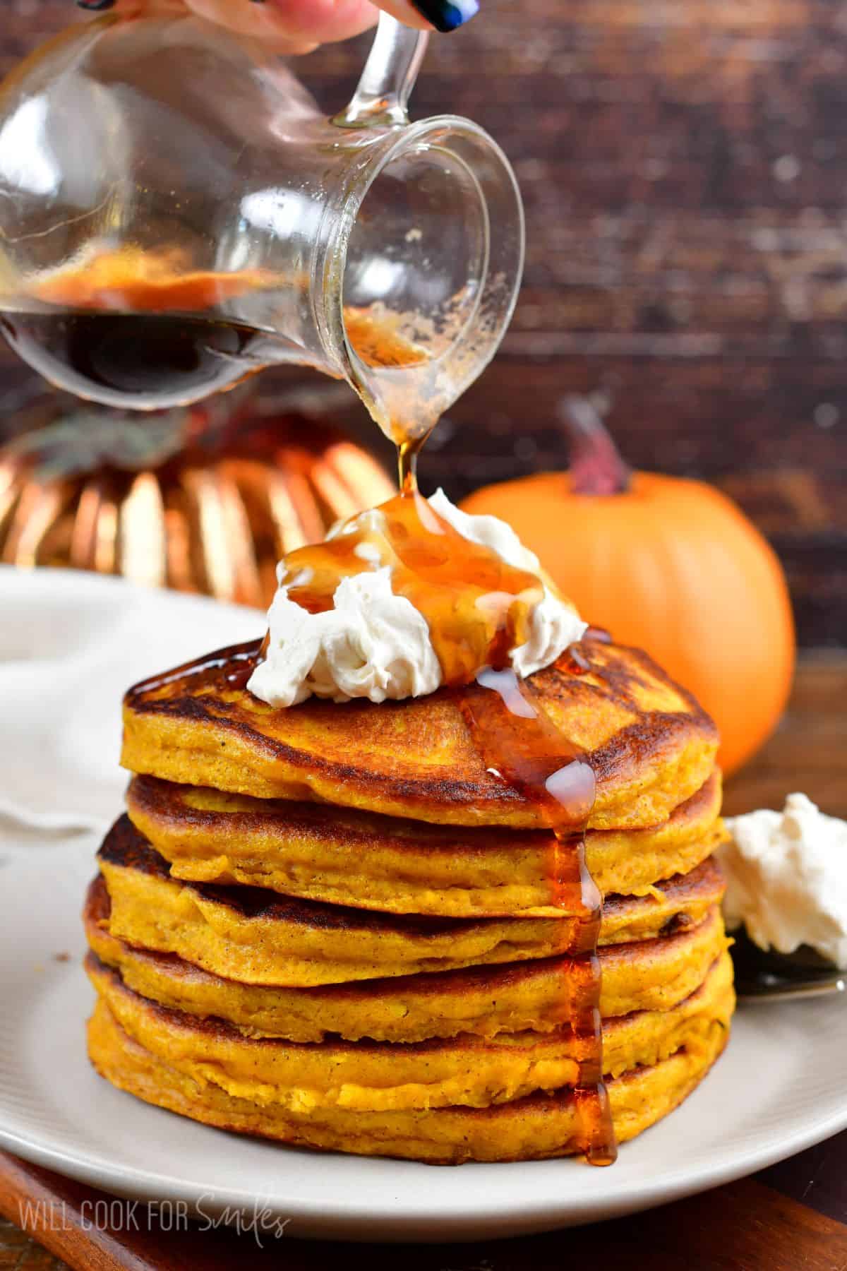 a stack of pumpkin pancakes on a plate with whipped cream on top and syrup being poured over the top.