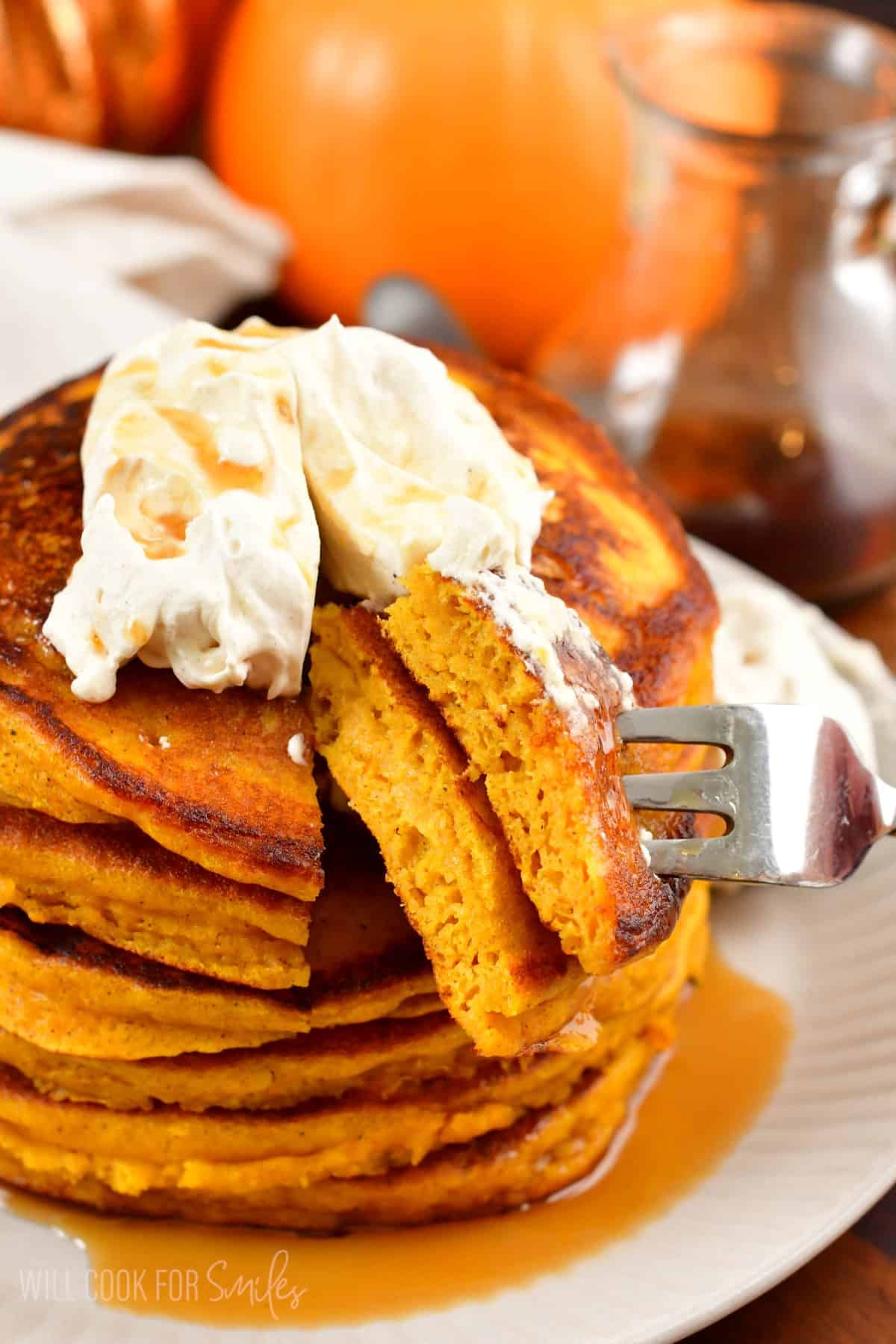A stack of pumpkin pancakes with whipped cream and syrup on top with a fork in taking some.