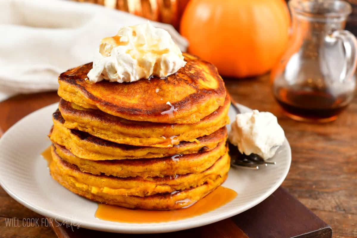 A stack of pumpkin pancakes on a plate with whipped cream on top and a spoon to the side with more whipped cream.