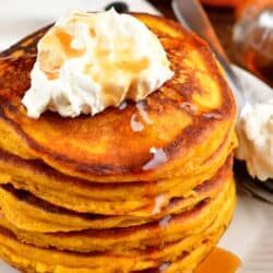 A stack of pumpkin pancakes with whipped cream and syrup on top on a plate.