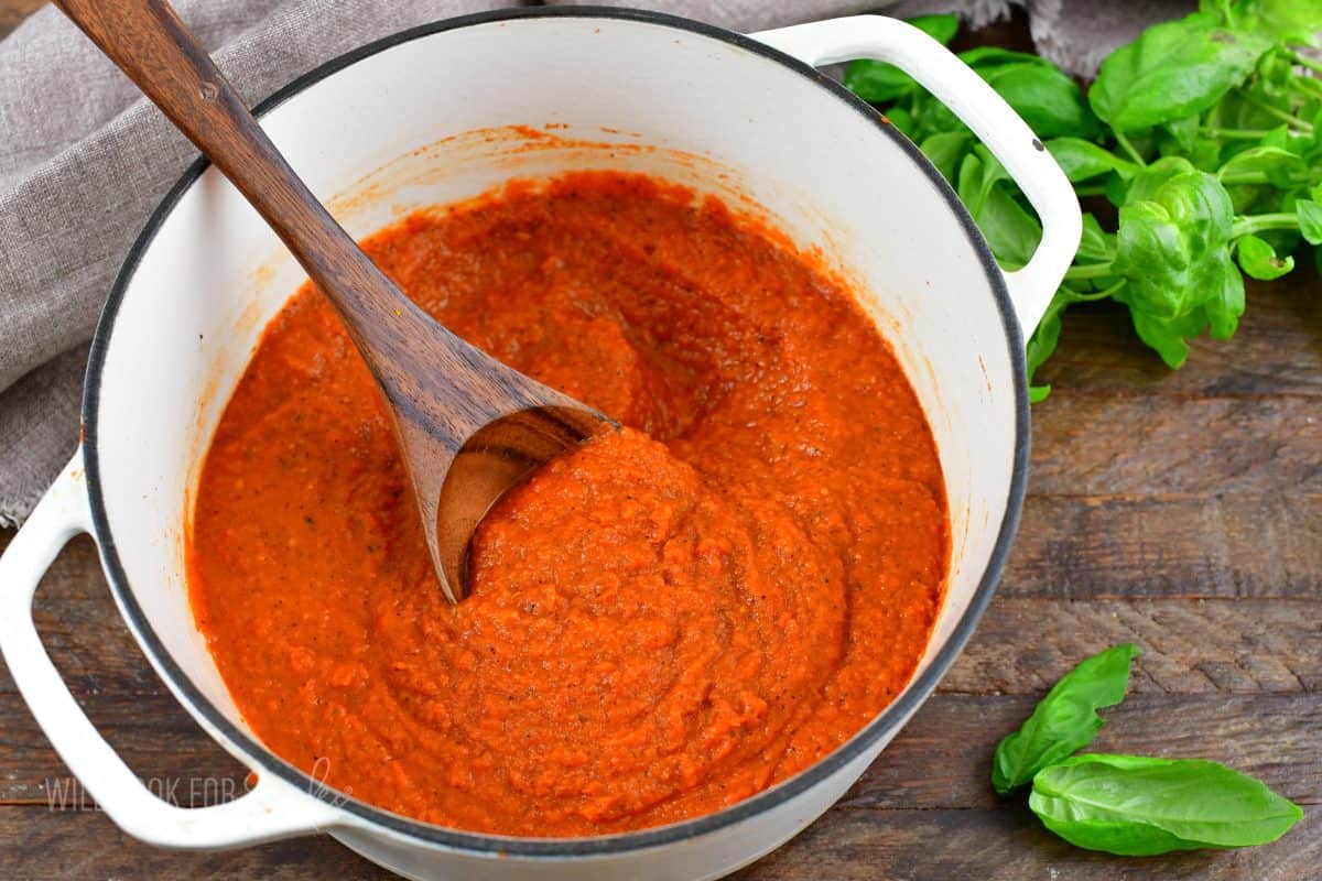 spaghetti sauce in a pot with a wooden spoon and basil on a wood surface.