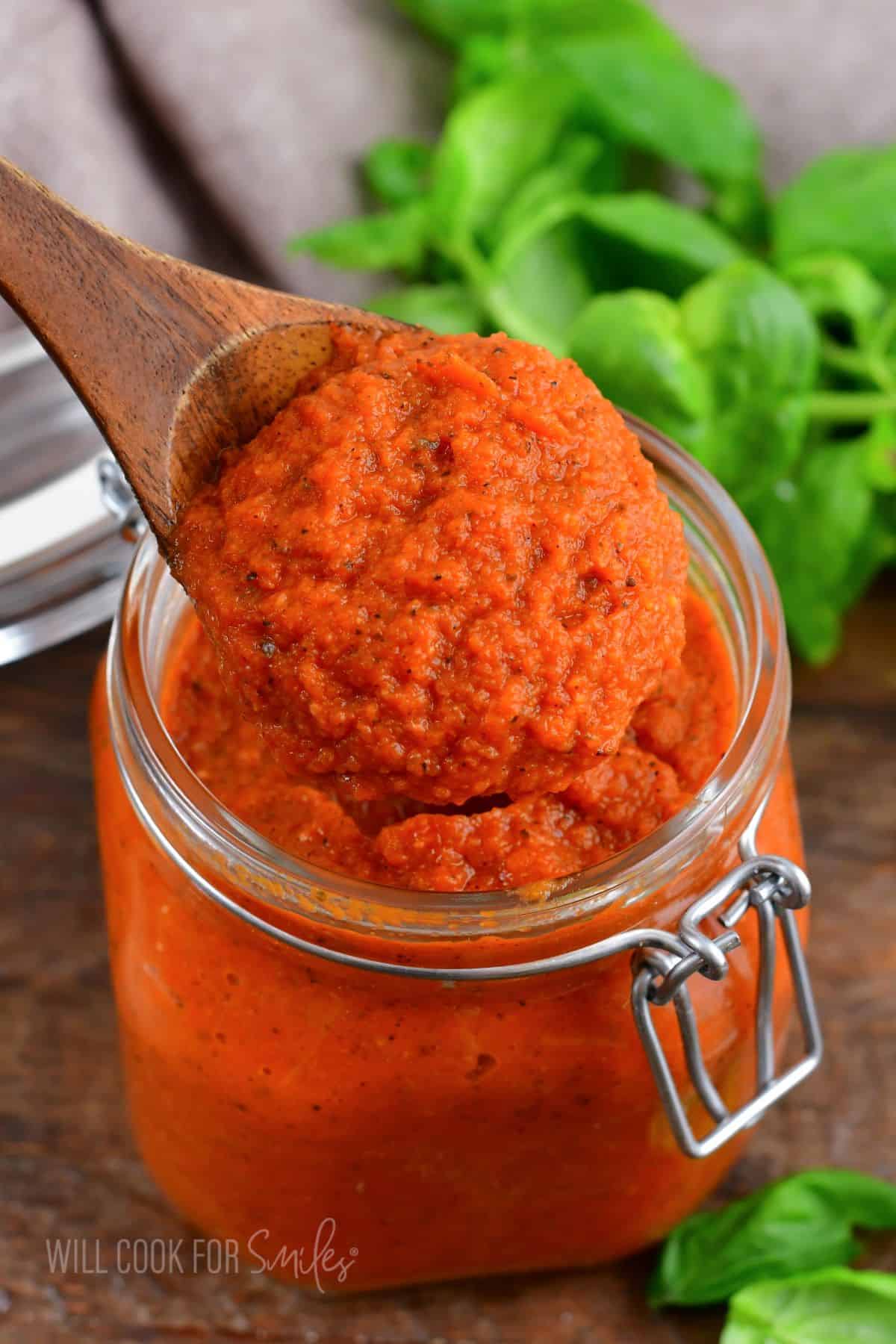 spaghetti sauce in a glass jar on a wooden surface with a wooden spoon lifting some out.