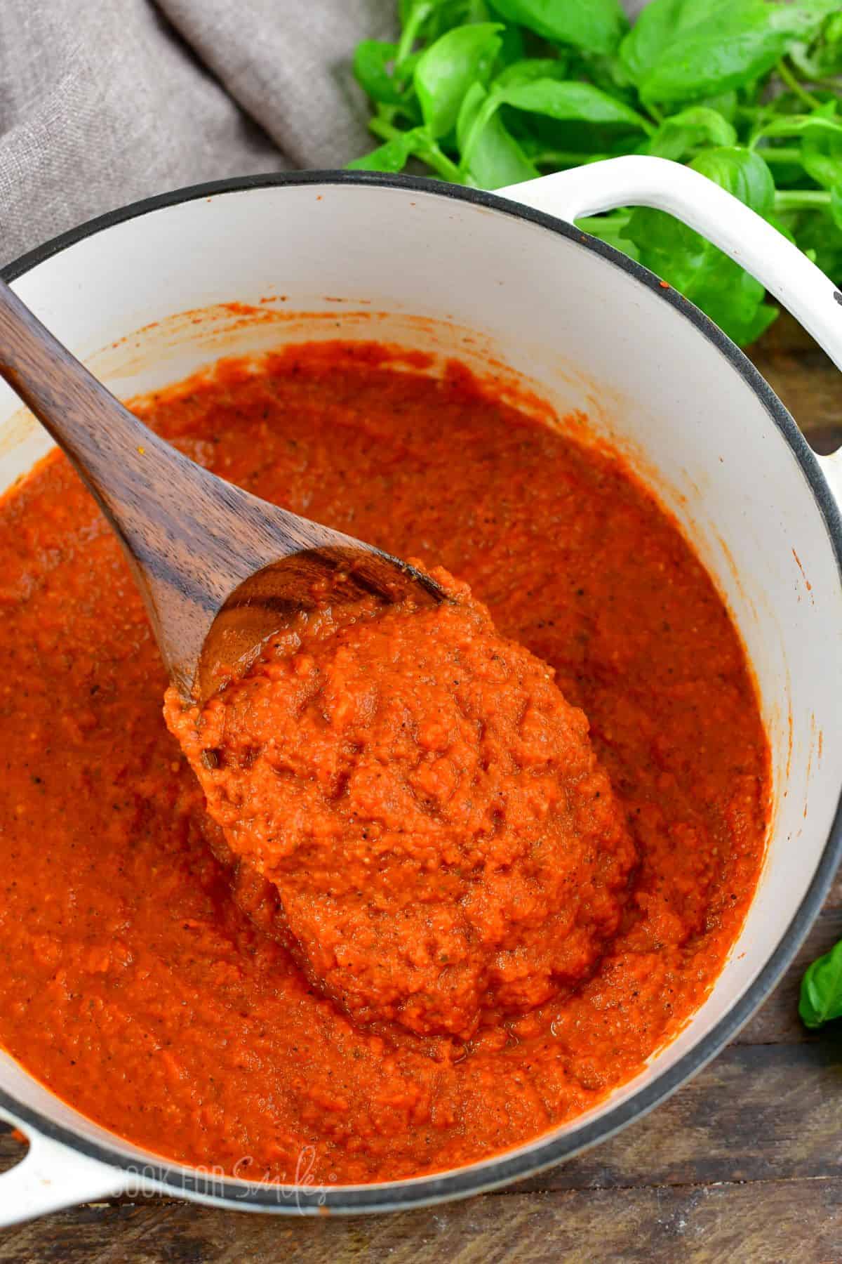 spaghetti sauce in a pot with a wooden spoon lifting sauce and basil on a wood surface.