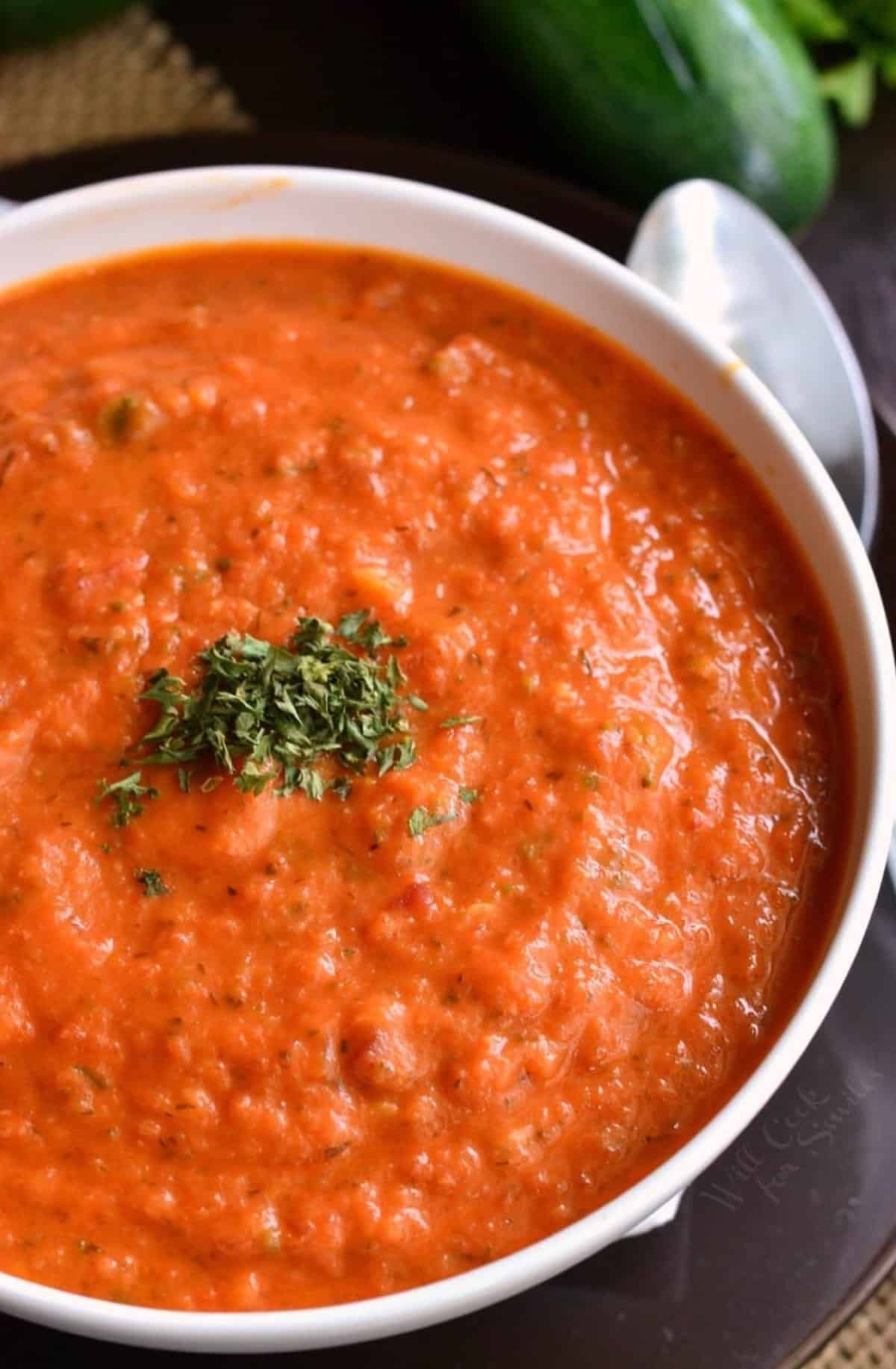 Tomato zucchini soup in a white bowl with a spoon to the side.