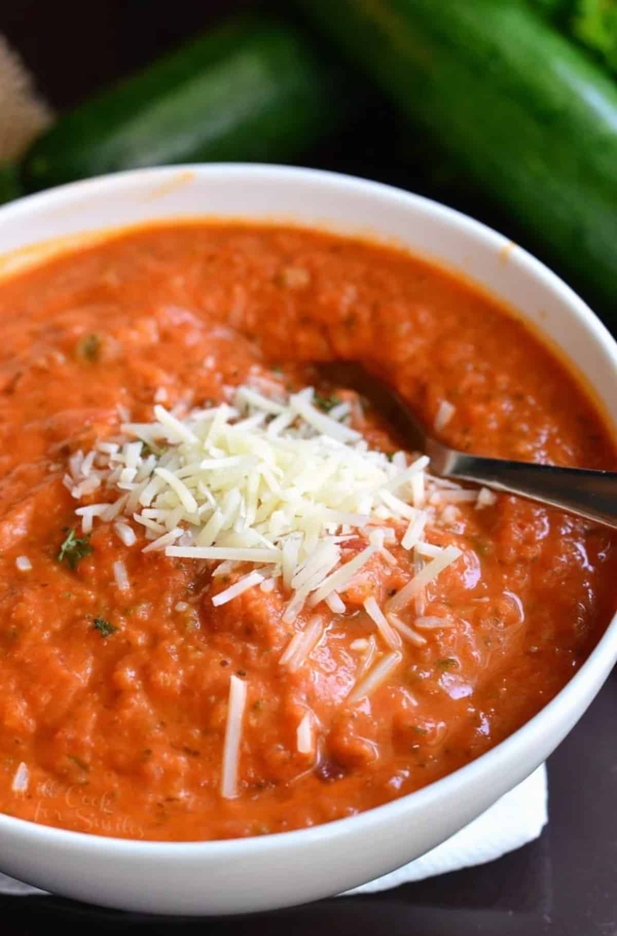 Tomato Zucchini soup in a white bowl with cheese on top as a garnish.