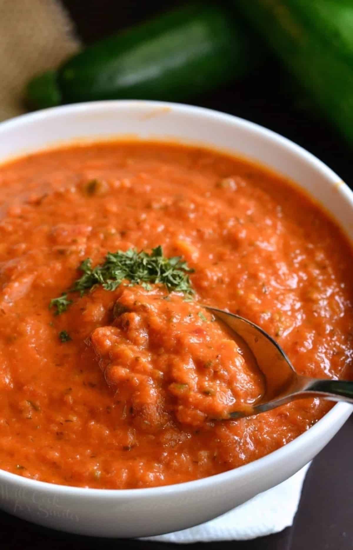 Tomato Zucchini soup in a white bowl with fresh basil and garnish on top and a spoon lifting some out.