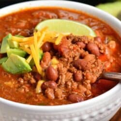 chili in a white bowl with a spoon in and cheese, avocado, and lime on top.