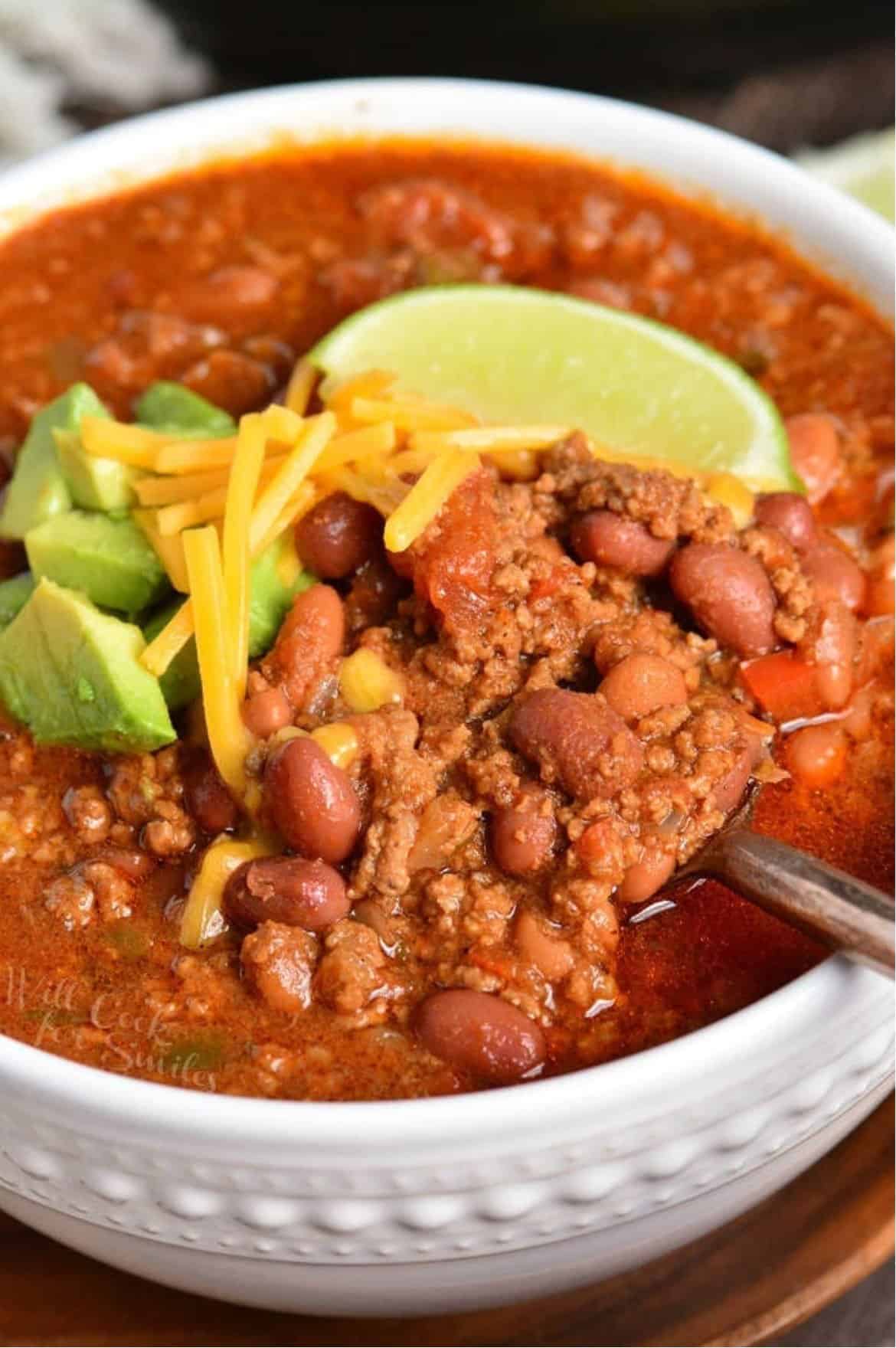 Bowl of chili with a spoon scooping some with avocado, cheese, and lime on top.