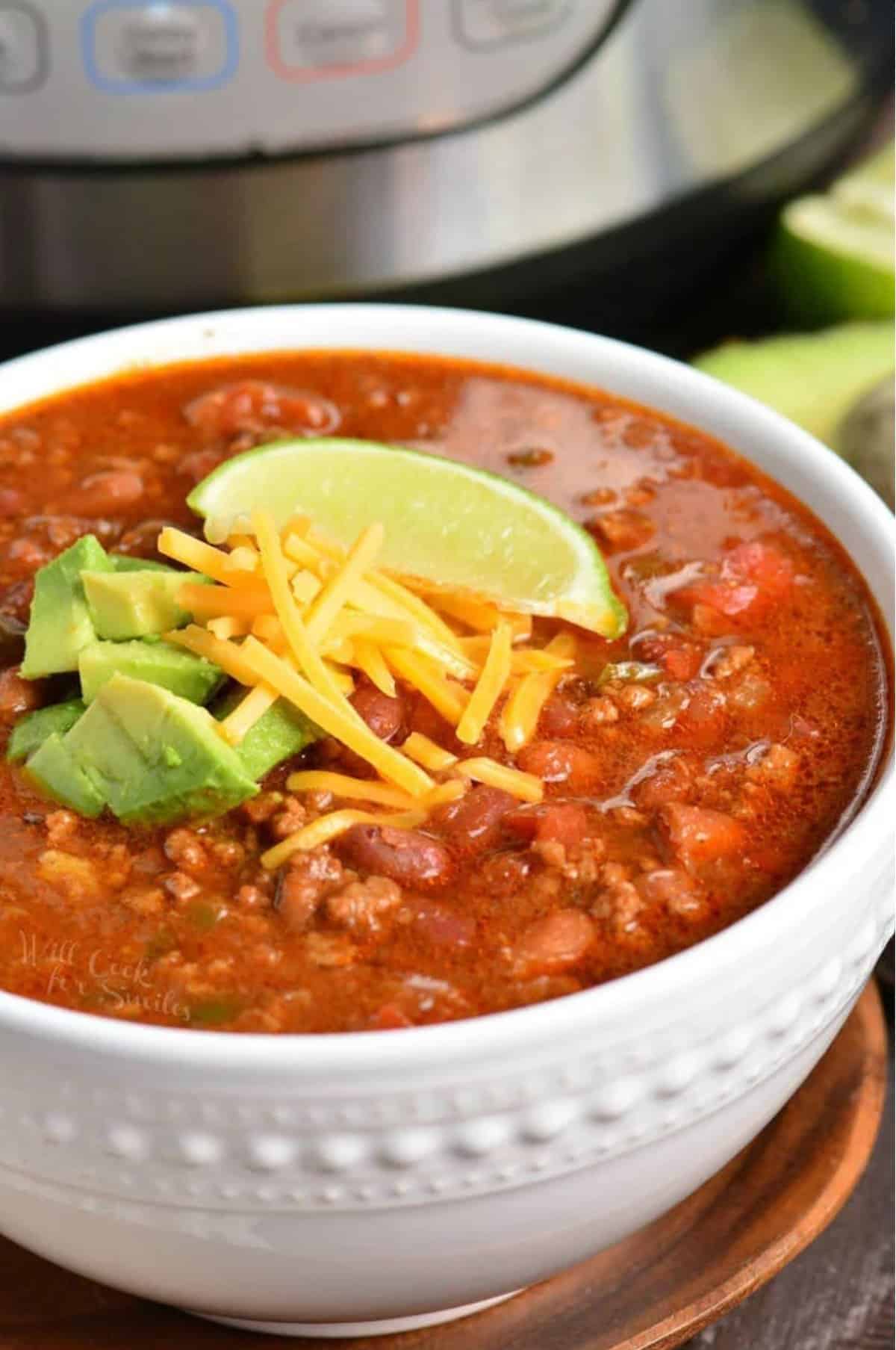 chili in a bowl on a wood plate with shredded cheese, avocado, and a lime wedge on top.