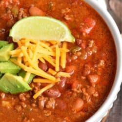 Chili in a bowl with shredded cheese, diced avocado, and a lime wedge on top.