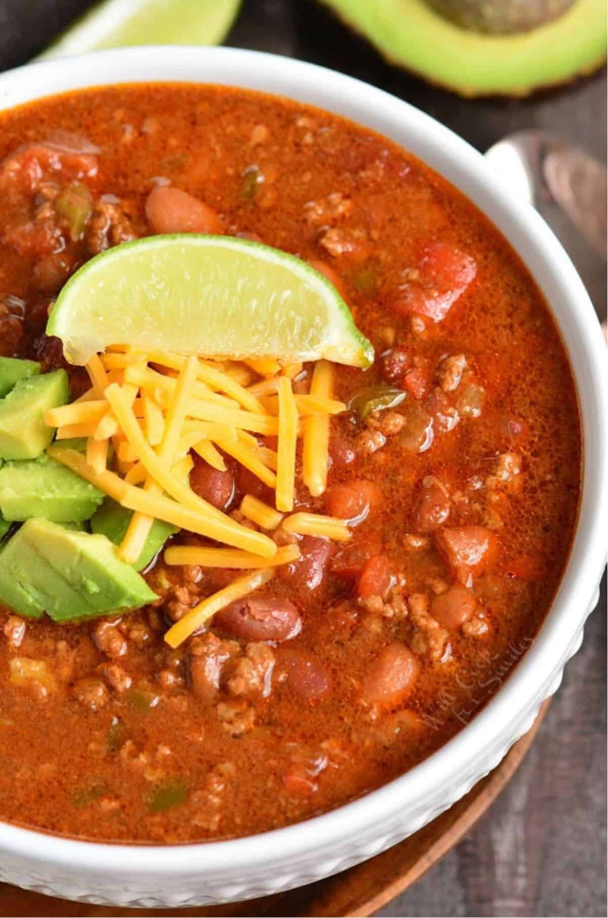 Chili in a bowl with shredded cheese, diced avocado, and a lime wedge on top as garnish.