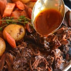 Pot roast, potatoes. carrots, in a bowl with a spoon pouring sauce over it.
