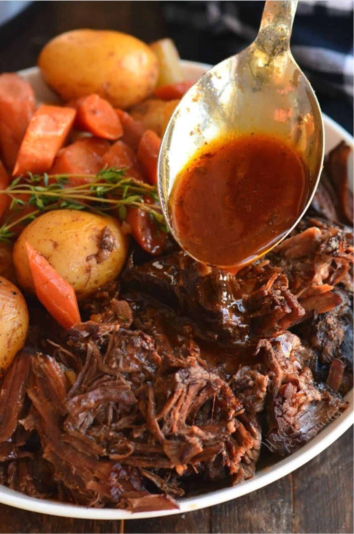 Pot roast, potatoes. carrots, in a bowl with a spoon pouring sauce over it.