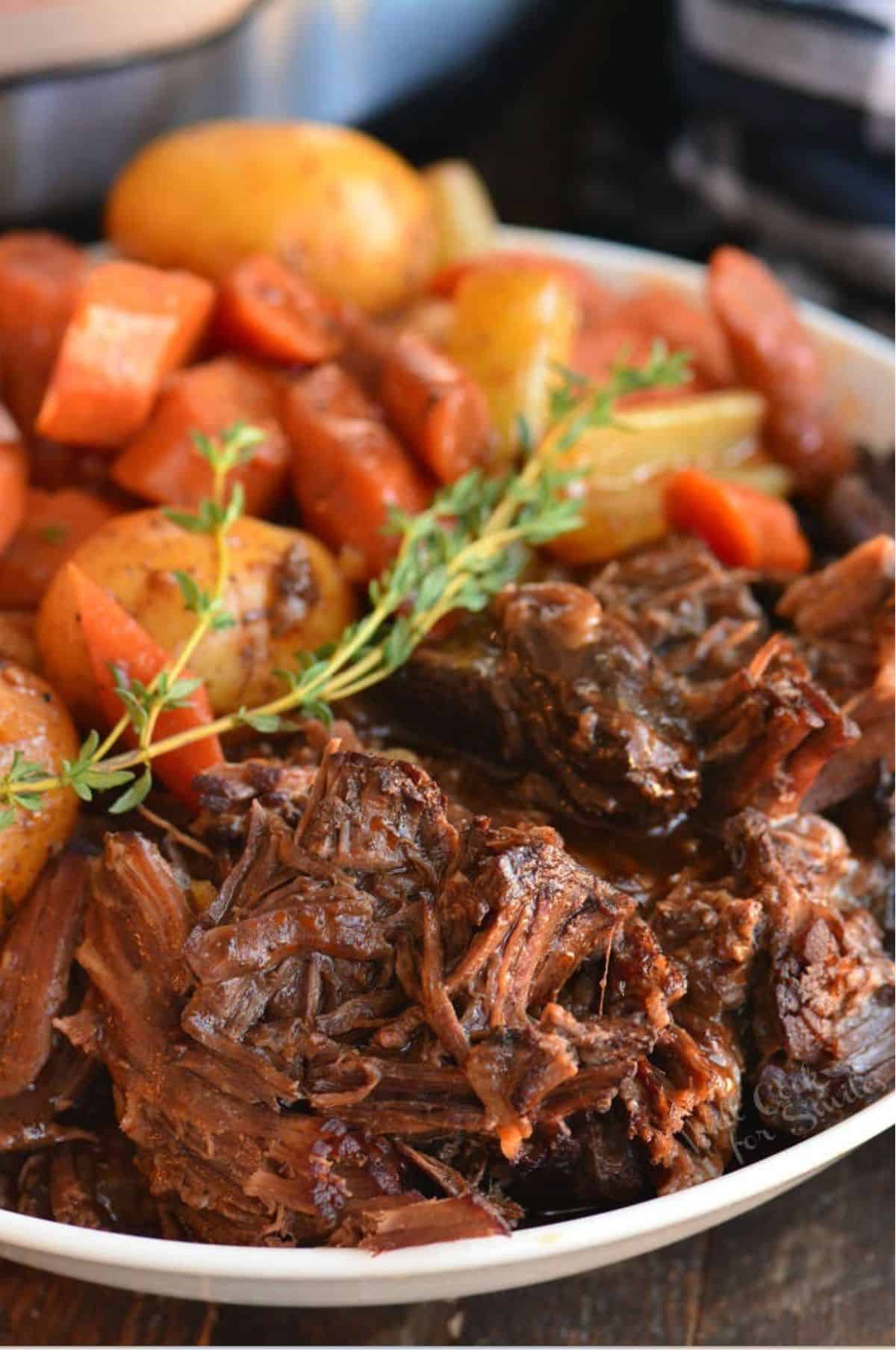 Pot roast with carrots and potatoes and a sprig of rosemary as garnish in a bowl.