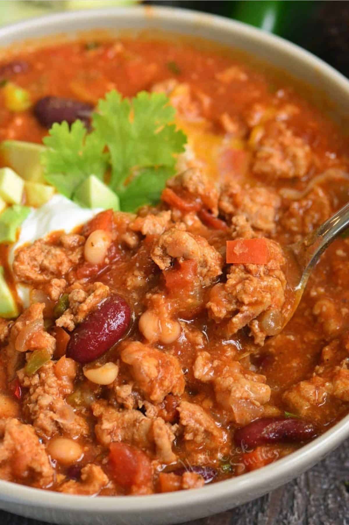 Chili in a bowl with a spoon and sour cream and diced avocado as garnish and a spoon scooping some chili.