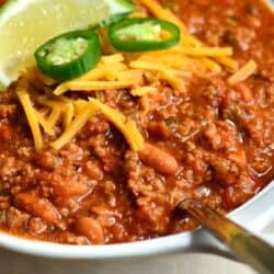 beef chili in the bowl with a spoon and topped with shredded cheddar, jalapeno slices, and lime.