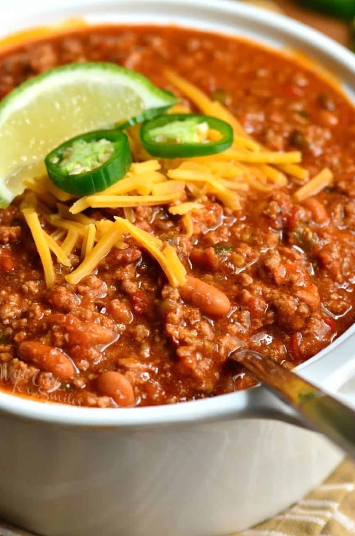 beef chili in the bowl with a spoon and topped with shredded cheddar, jalapeno slices, and lime.