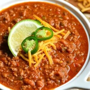 beef chili in a white serving pot with jalapeno, cheese, and lime garnishes.