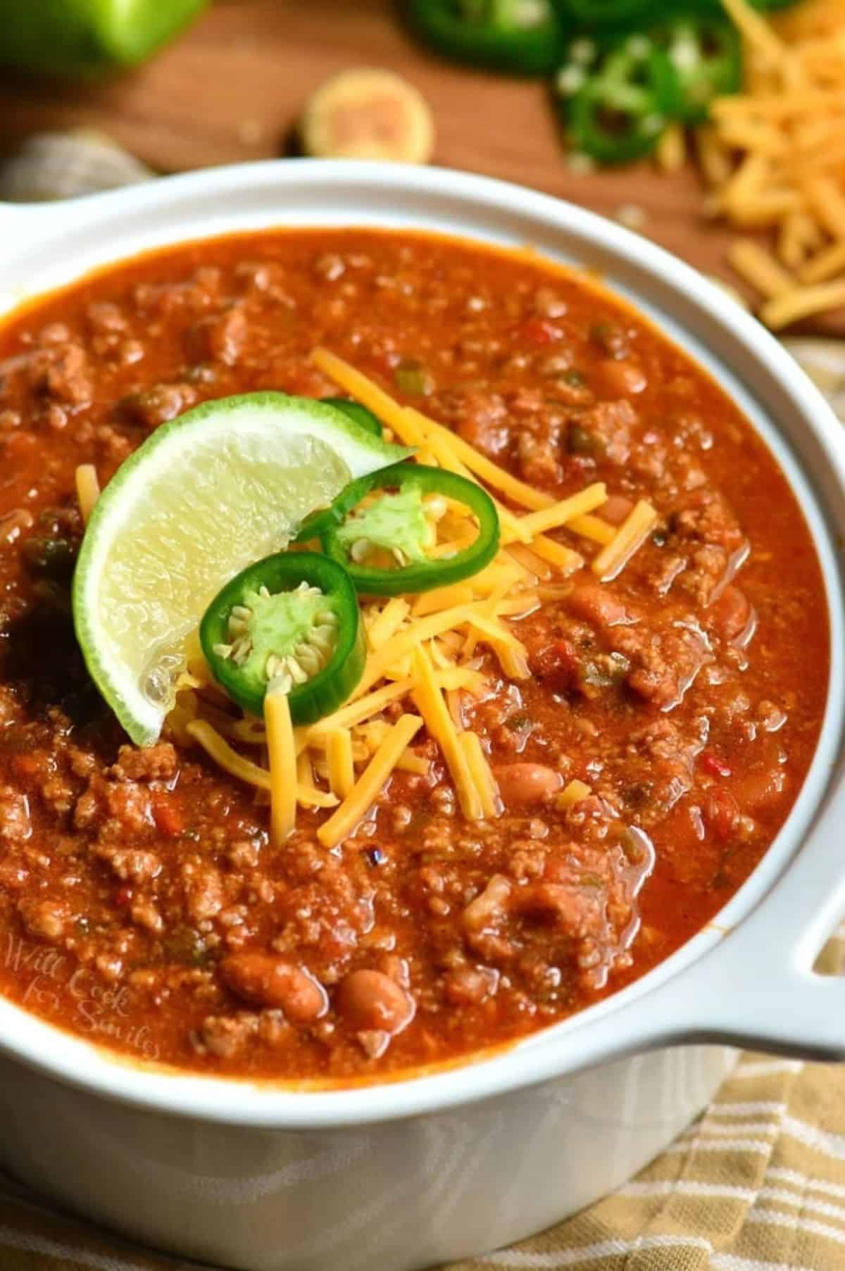 beef chili in a white serving pot with jalapeno, cheese, and lime garnishes.