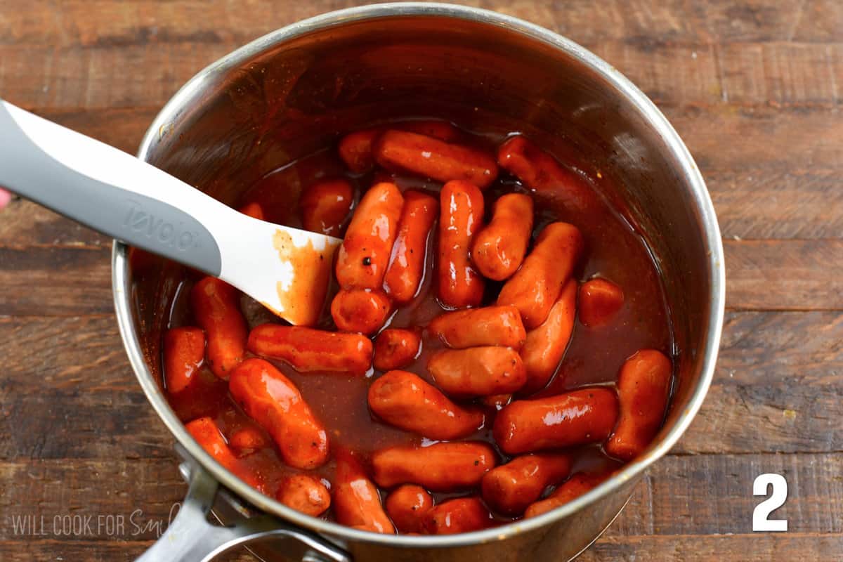 BBQ sauce and little smokies in a pot with a plastic spoon on a wood surface.