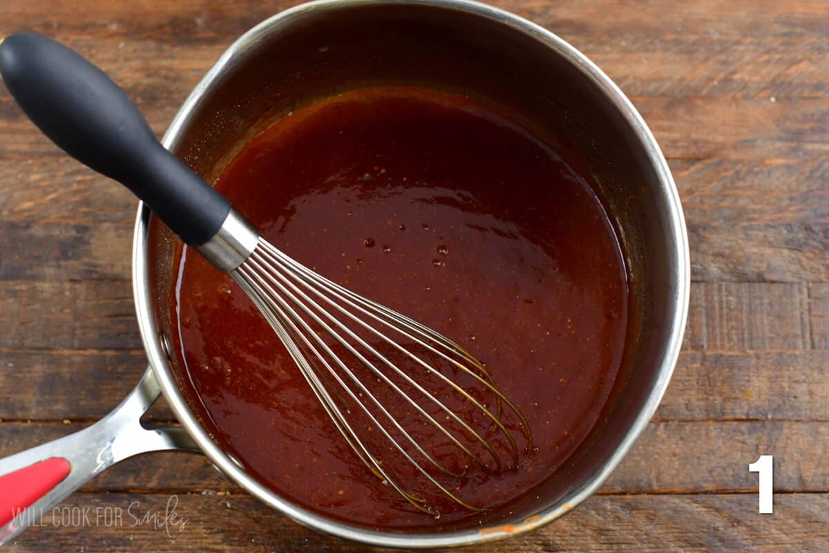 BBQ Sauce in a pot with a metal whisk in the pot on a wood surface.
