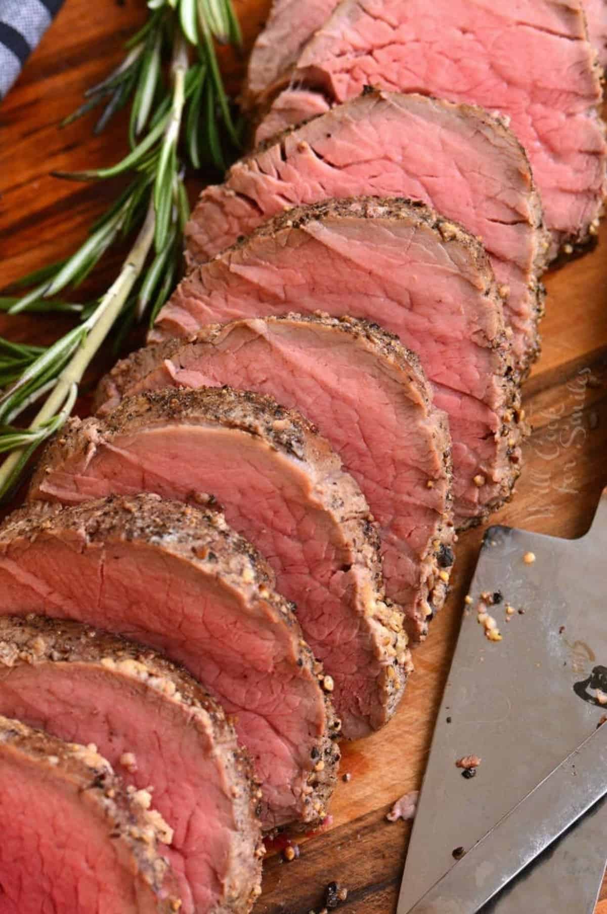 sliced beef tenderloin with seasoning on the sides and rosemary sprig next to it.